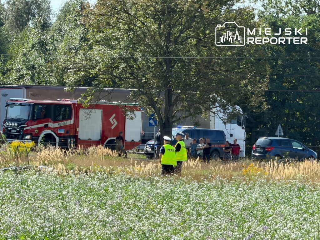 Na miejscu zdarzenia stoi czerwony wóz strażacki, a obok grupka osób obserwujących działania służb ratunkowych.