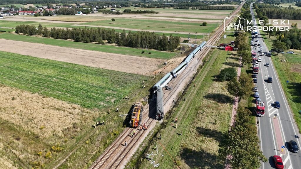Wielu ratowników pracuje przy wykolejonym pociągu na torach, obok widać stojące samochody i służby ratunkowe.