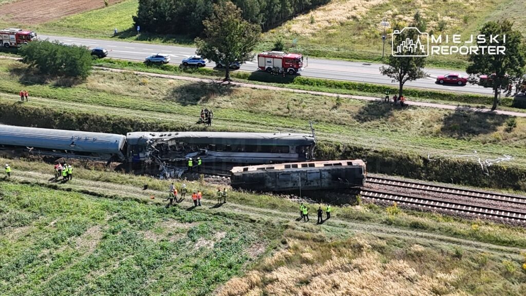Złamanie torów kolejowych po katastrofie, z uszkodzonym pociągiem i ratownikami w kamizelkach na miejscu zdarzenia.