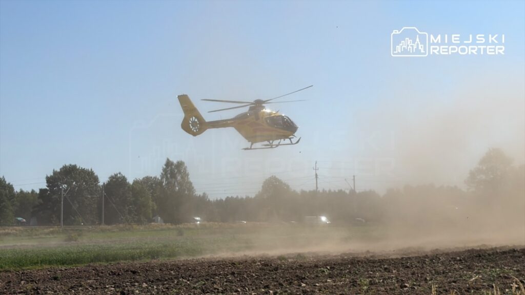Żółty helikopter wznosi się nad pola, unosząc chmurę kurzu w słoneczny dzień.