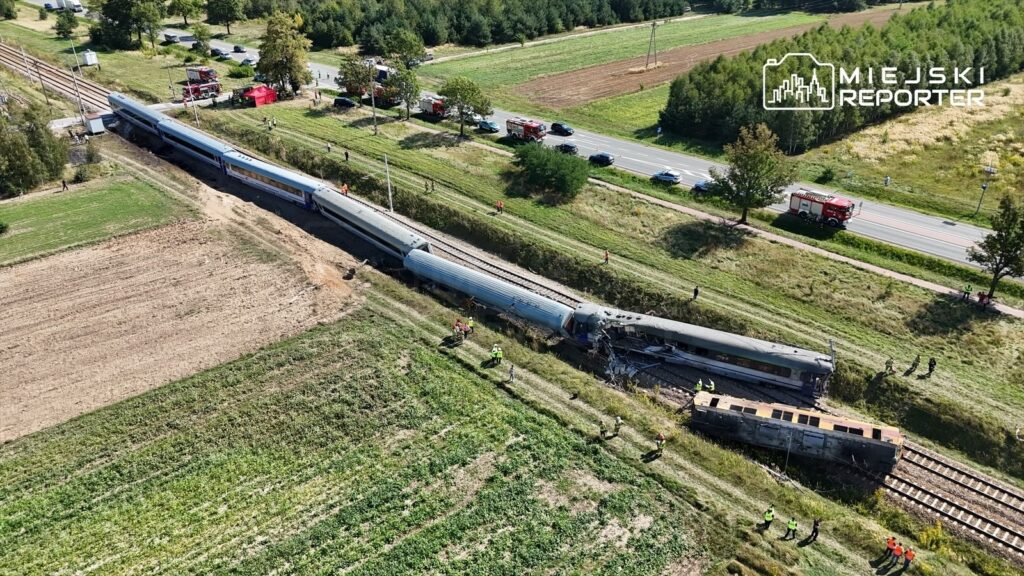 Złomowany pociąg leży na torach w otoczeniu ratowników i służb emergency, w tle widoczne pola i droga.