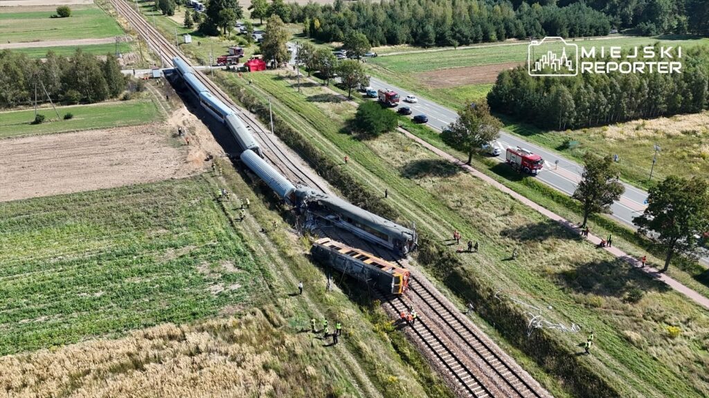 Zderzenie pociągu z ciężarówką na torach; na miejscu pracują służby ratunkowe, a w tle widać zniszczony wagon.