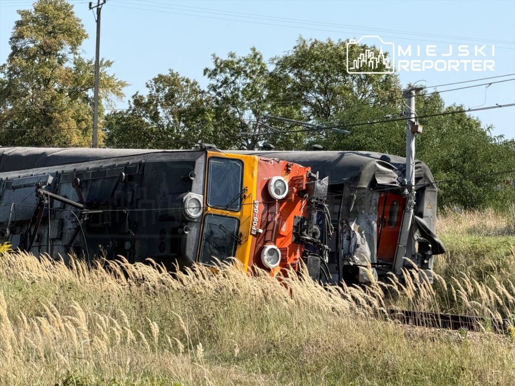 Złamany pociąg leży na boku wśród wysokiej trawy, z uszkodzoną kabiną i widocznymi zniszczeniami.