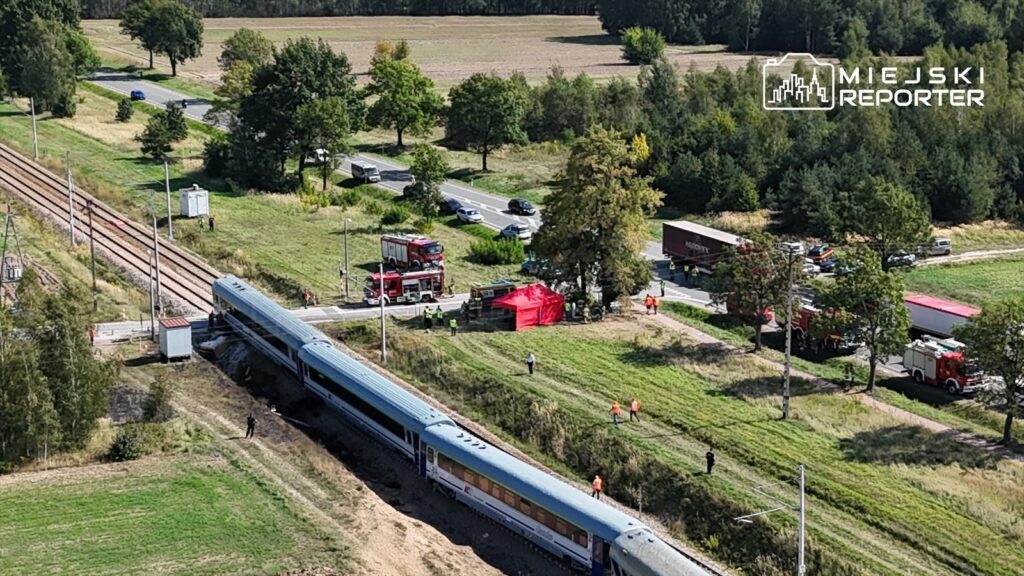 Pociąg leży na boku obok torów kolejowych, a w tle widać służby ratunkowe oraz pojazdy na drodze.