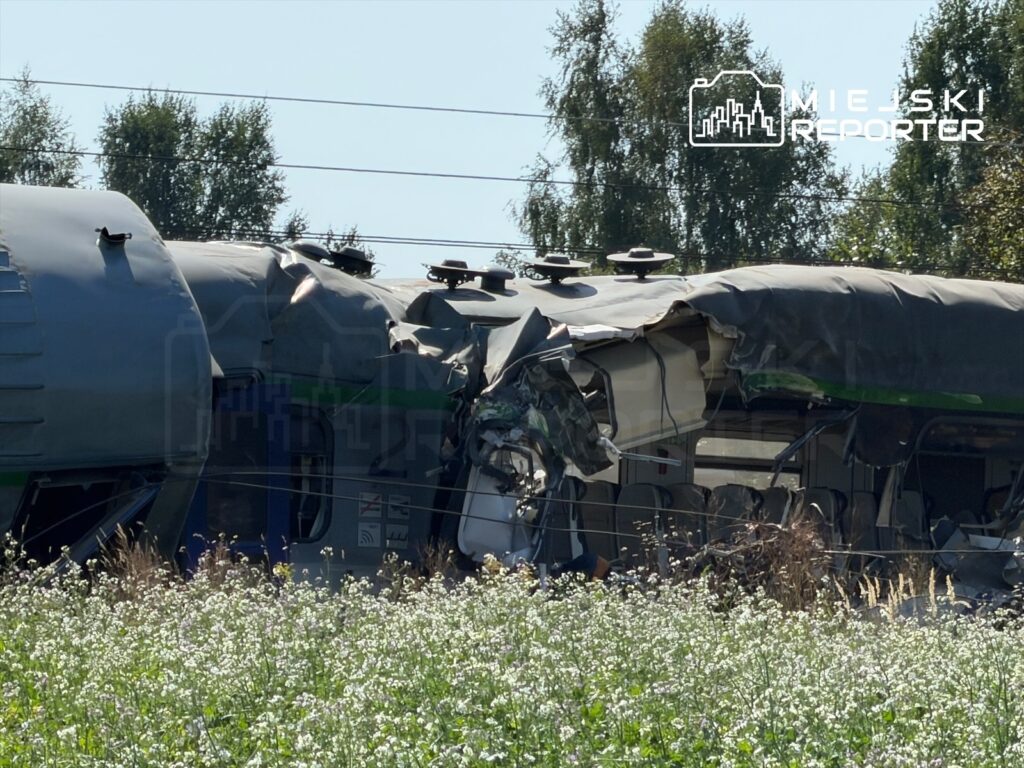 Zniszczony wagon pociągu leży w polu, otoczony kwiatami, z widocznymi uszkodzeniami na dachu i ścianach.