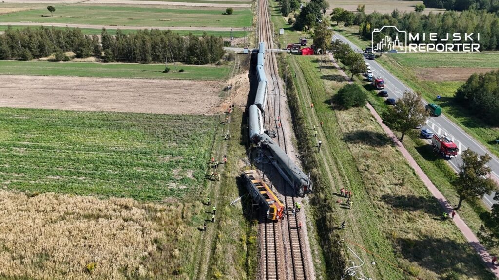 Wagon kolejowy wykolejony na torach, obok pracują ratownicy w odblaskowych kamizelkach, w tle zielone pola.
