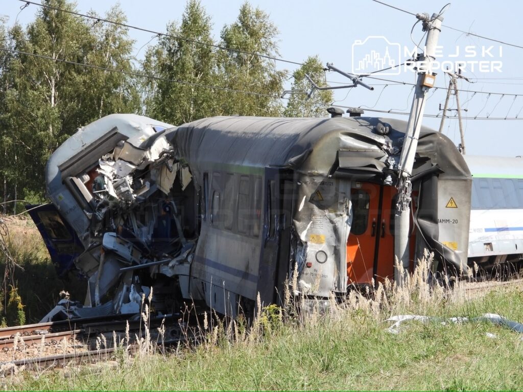 Zderzony pociąg z uszkodzoną kabiną maszynisty leży na torach w otoczeniu zieleni i drzew.