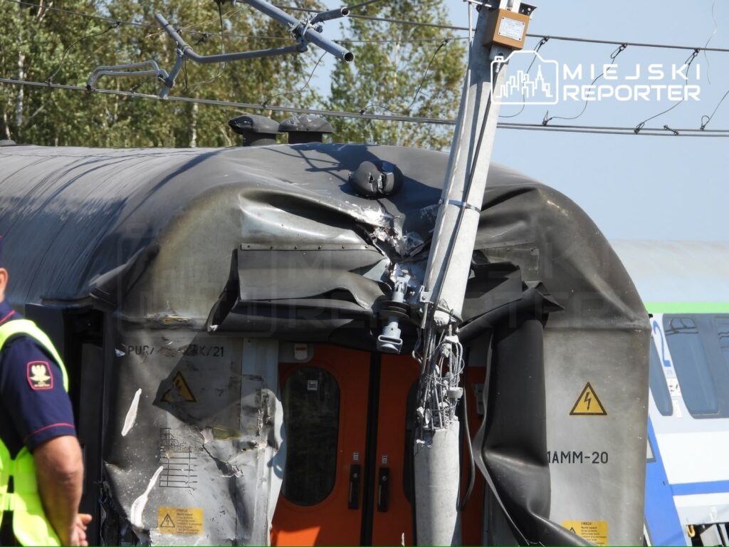 Zniszczony wagon kolejowy z uszkodzoną dachową częścią oraz przewodami elektrycznymi, obok stoi policjant w kamizelce.