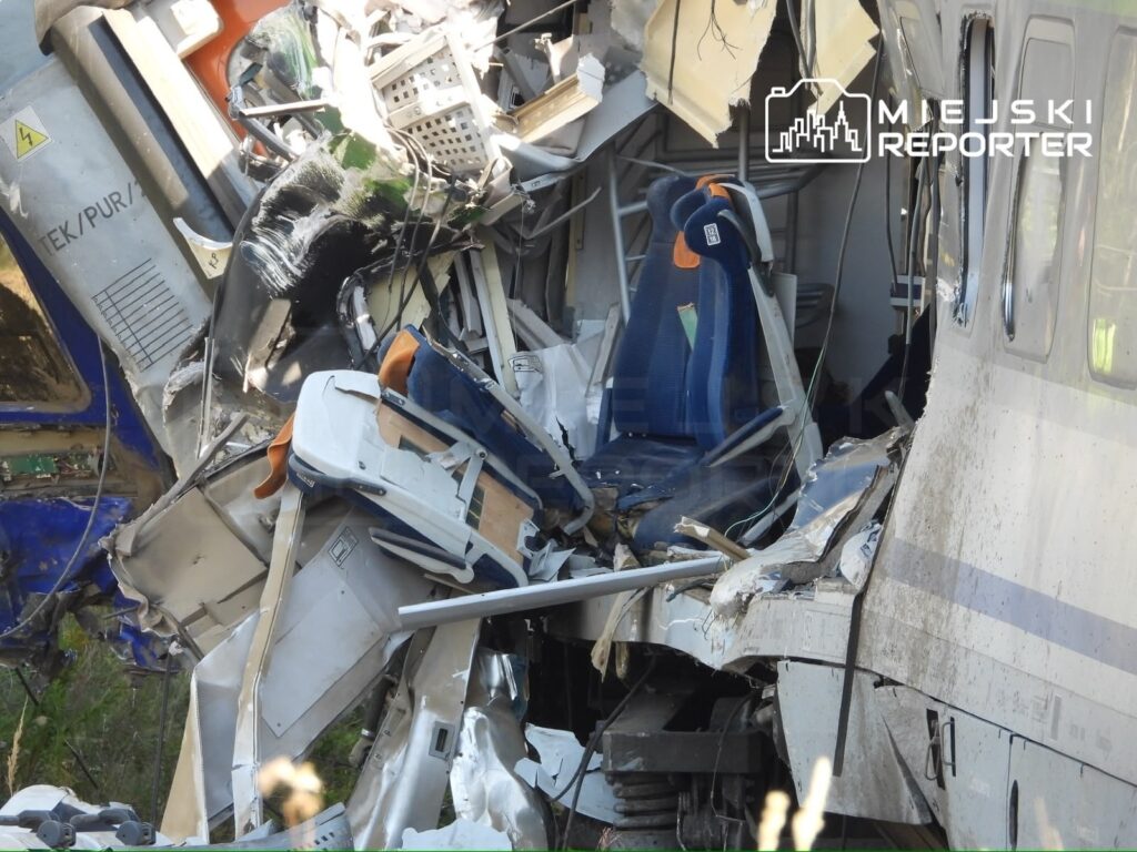 Zniszczony wagon kolejowy z uszkodzonymi siedzeniami i rozbitymi elementami wnętrza, otoczony roślinnością.