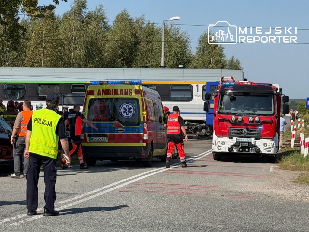 Na drodze, w pobliżu torów kolejowych, zespół ratunkowy i policja interweniują przy wypadku. W tle stoi pociąg.