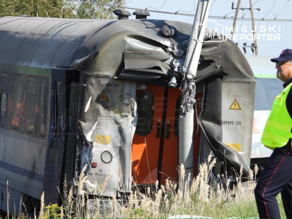 Zniszczony wagon kolejowy z uszkodzoną przednią częścią, obok którego stoi mężczyzna w kamizelce odblaskowej.