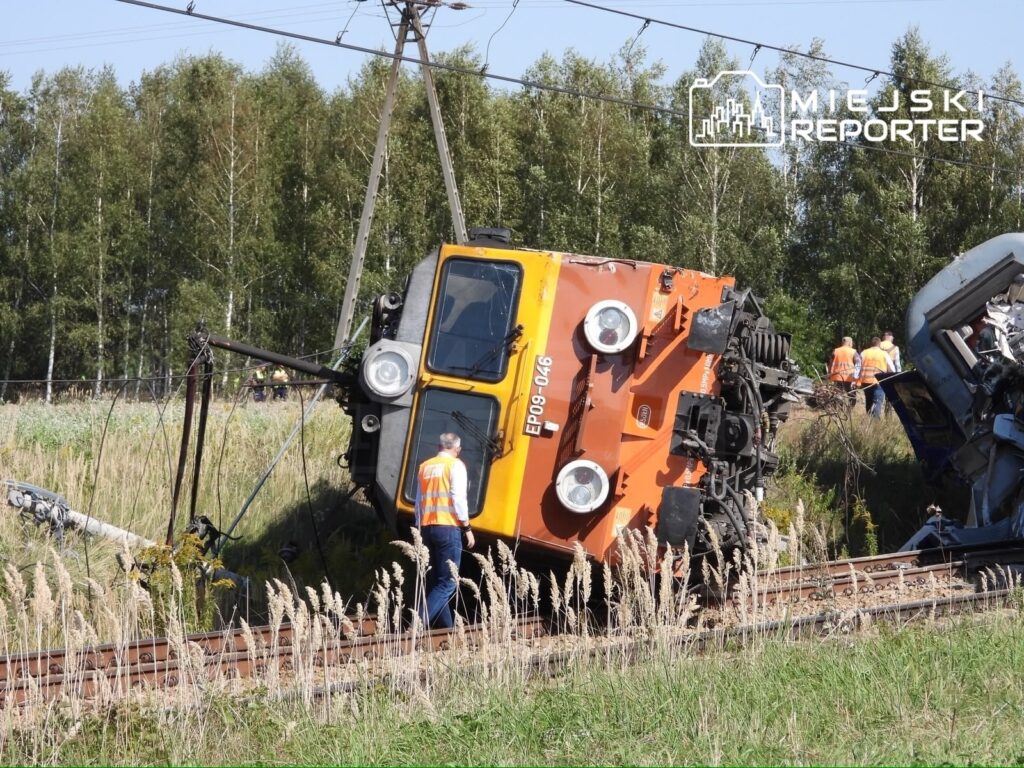 Złomowany pojazd kolejowy przewrócony na torach, obok pracownicy w kamizelkach odblaskowych.