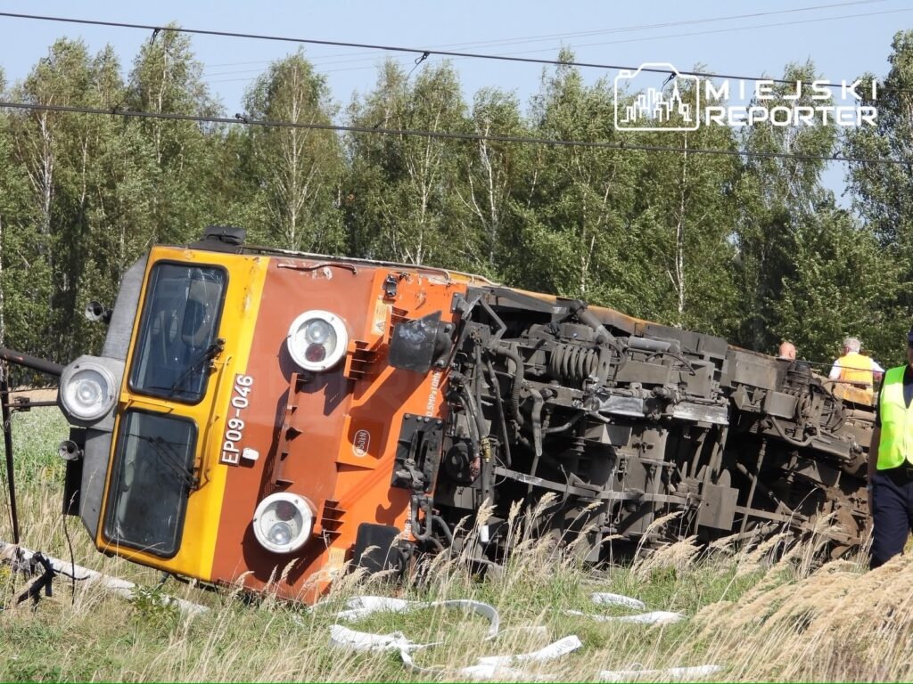 Złomowany pociąg leżący na boku wśród trawy, obok pracownicy w odblaskowych kamizelkach.
