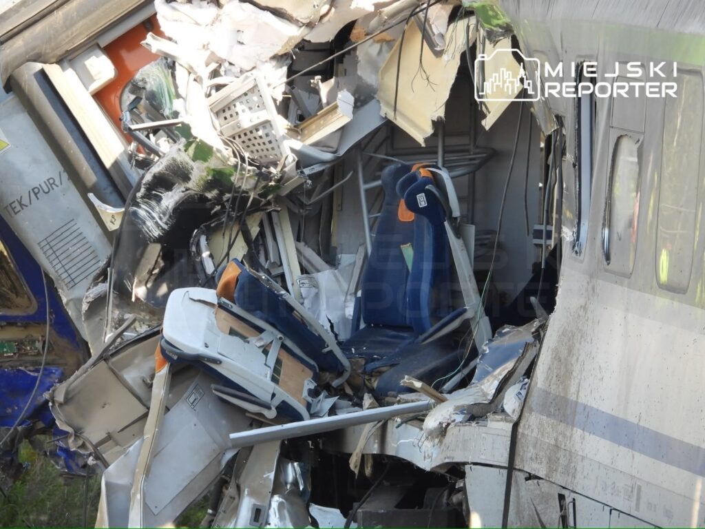Zniszczony wagon kolejowy z wywróconymi siedzeniami i uszkodzoną konstrukcją, leżący na boku.
