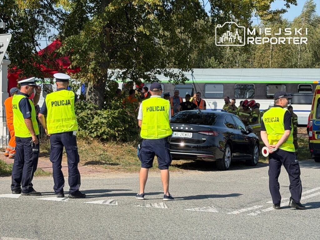 Grupa policjantów w kamizelkach odblaskowych obserwuje działania ratunkowe przy drodze, obok czarnego samochodu.