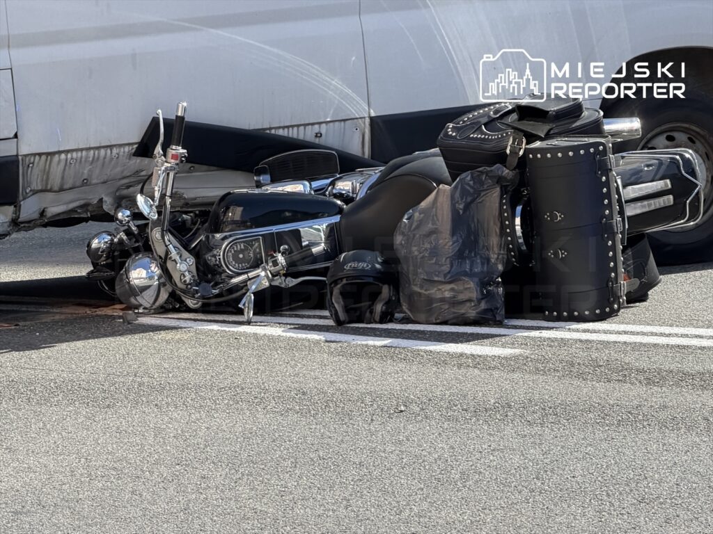 Motocykl leżący na boku na jezdni, w pobliżu białego vanu, z widocznymi uszkodzeniami.