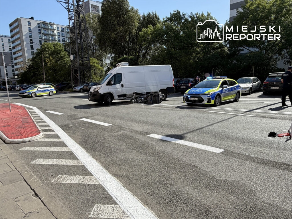 Motocyklista leży na jezdni obok przewróconego motocykla, a policjanci zabezpieczają miejsce wypadku w miejskim otoczeniu.
