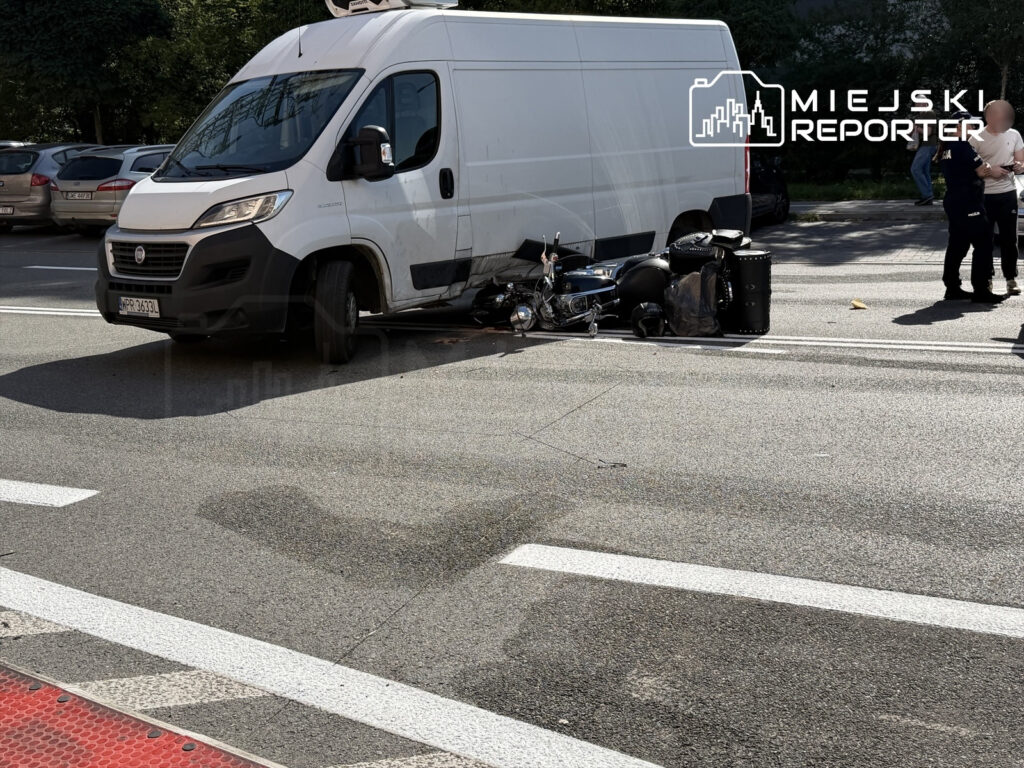Motocyklista leży obok przewróconego motocykla, a obok stoi biały furgon. W tle widać zaparkowane samochody.