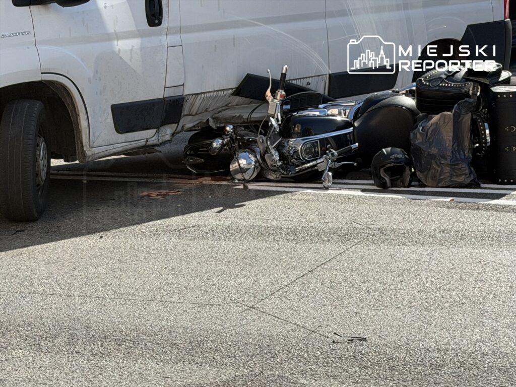 Motocykl leży na ziemi obok białego furgonu, na drodze w miejskim otoczeniu, widoczne plamy krwi.