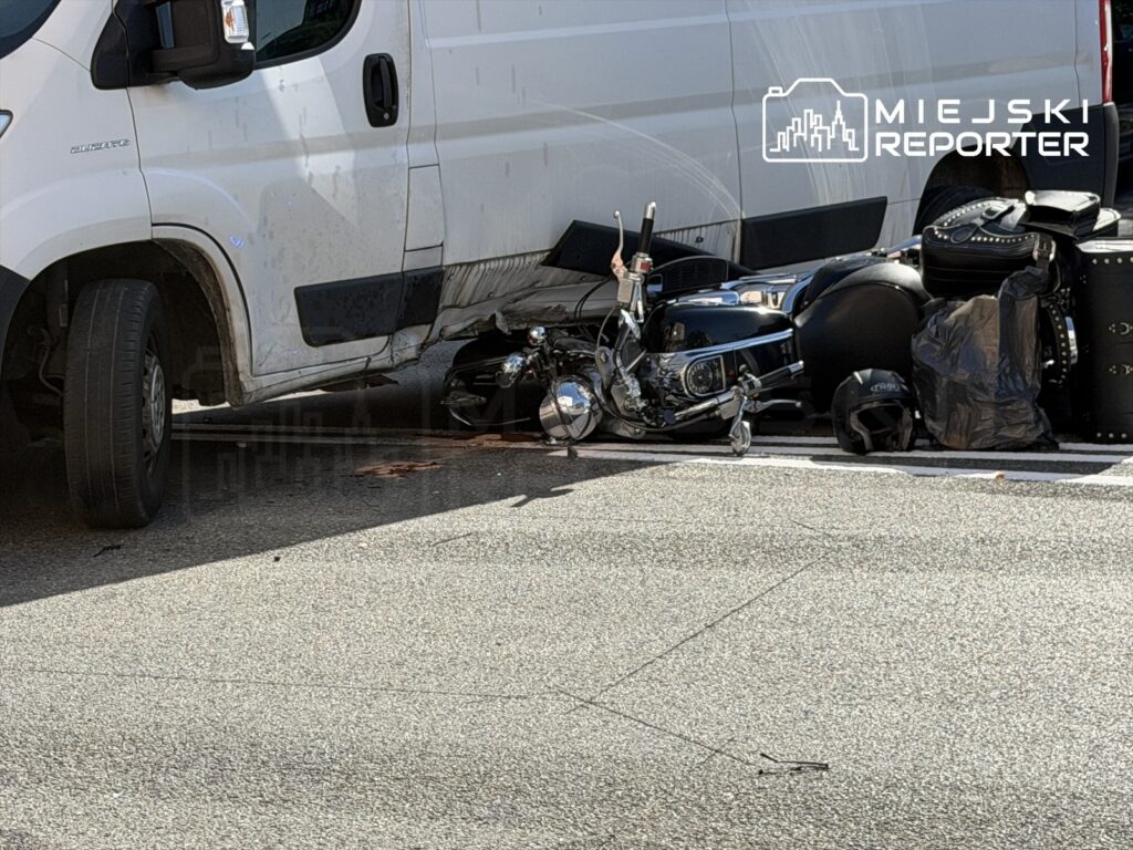 Motocykl leżący na jezdni obok białego furgonu, z widocznymi uszkodzeniami i akcesoriami motocyklowymi w tle.