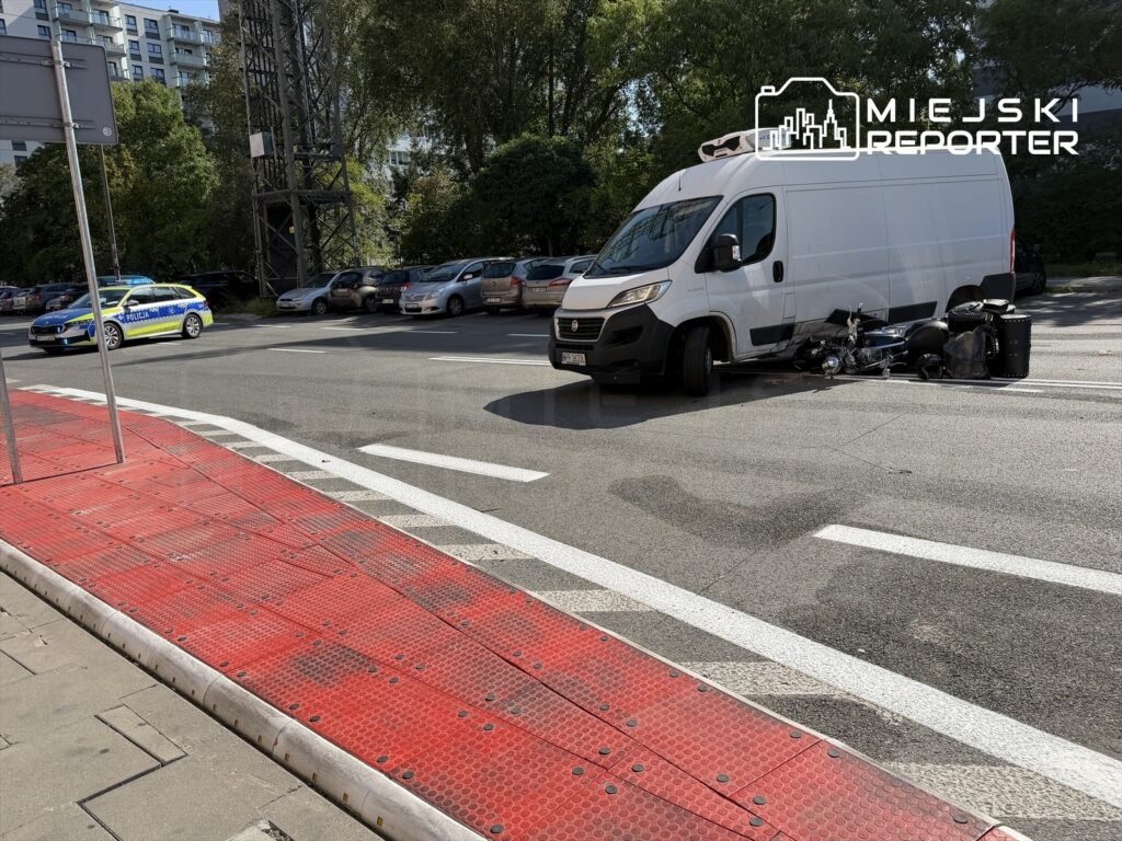 Biały furgon zatrzymany na drodze, obok leży przewrócony motocykl oraz policyjny radiowóz w tle.