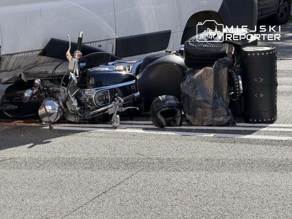 Motocykl leżący na jezdni obok czarnego plecaka i kasku, w tle widoczny biały samochód dostawczy.