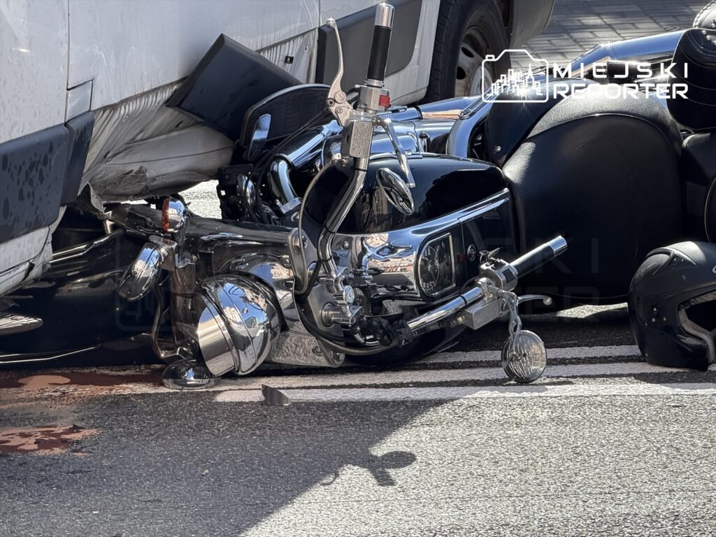 Motocykl leżący na jezdni, obok widoczna plama krwi, w tle częściowo widoczny biały pojazd.