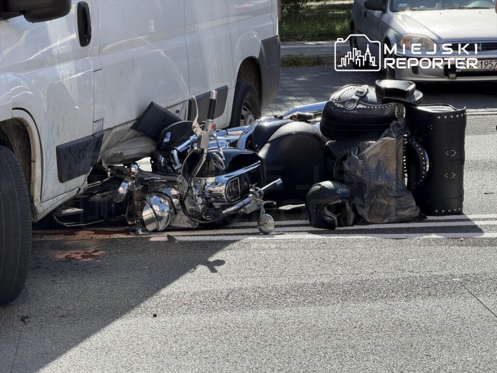 Motocykl leży na jezdni obok białego furgonu, z widocznymi uszkodzeniami i plamą krwi na asfalcie.