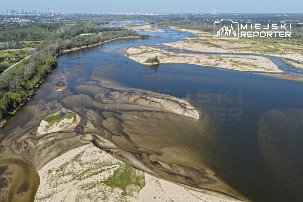 Szeroka panorama rzeki z odsłoniętymi piaszczystymi brzegami i wyspami, w tle zarys nowoczesnych budynków.