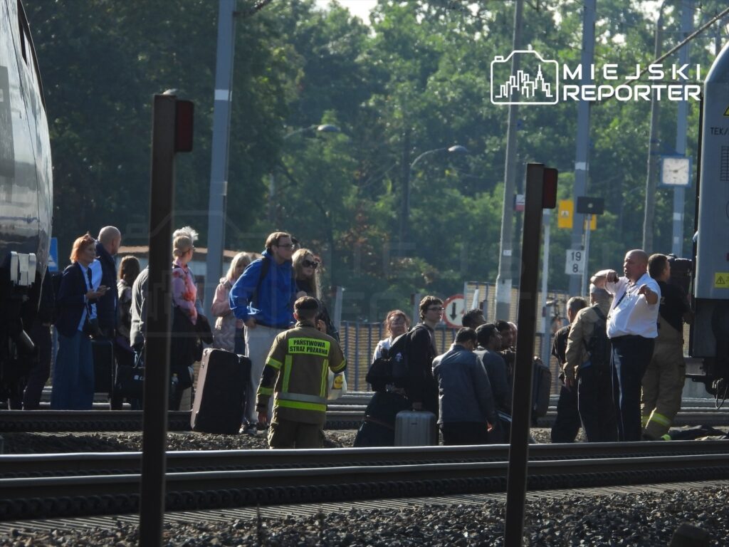 Grupa pasażerów z bagażami stoi na peronie obok pociągu, podczas gdy strażak rozmawia z pracownikiem kolei.