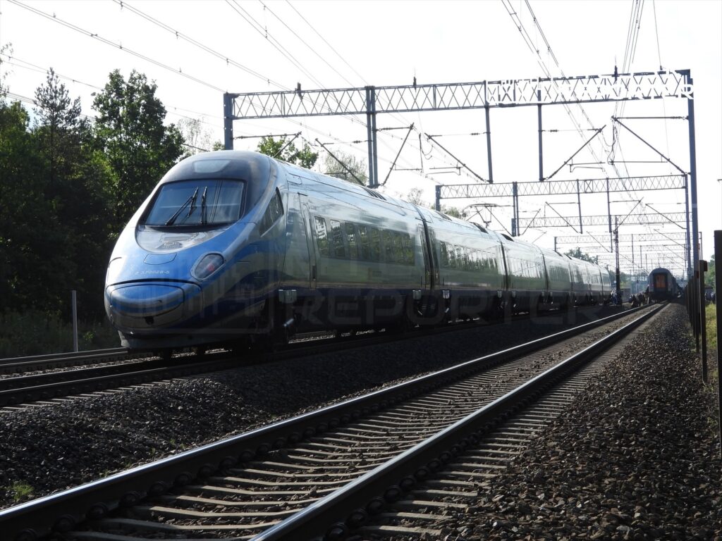 Pociąg Pendolino poruszający się po torach w otoczeniu zieleni, z widocznymi słupami trakcyjnymi w tle.