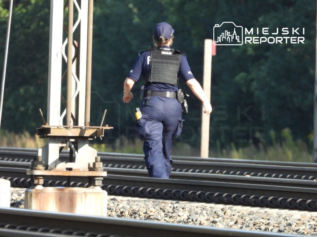 Policjant w kamizelce taktycznej idzie wzdłuż torów kolejowych, monitorując sytuację po wypadku z mężczyzną.