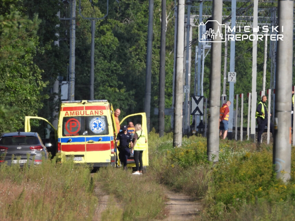 Pojazd ratunkowy na torach kolejowych, obok grupy osób w odzieży roboczej, w otoczeniu zieleni i słupów elektrycznych.