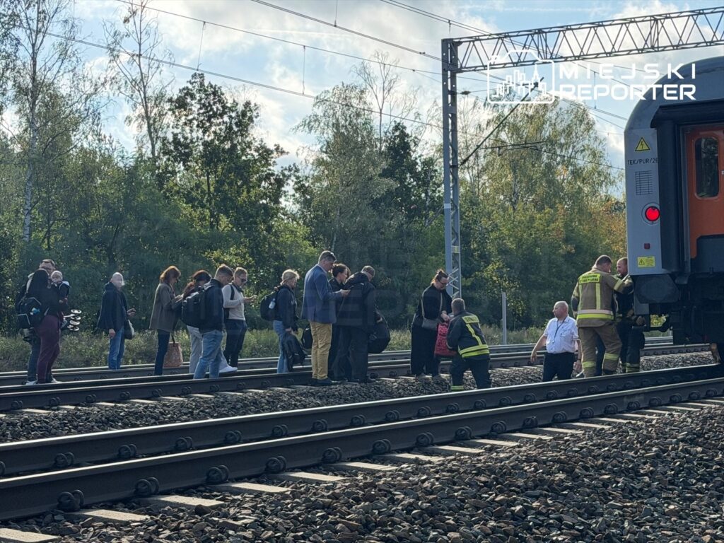 Grupa ludzi, w tym mężczyzn, stoi na torach kolejowych obok pociągu, a służby ratunkowe udzielają pomocy.