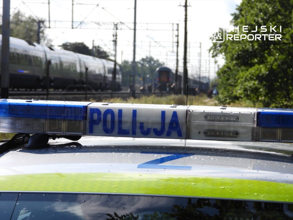 Policyjny radiowóz z niebiesko-białym sygnalizatorem stoi na torach kolejowych, w tle przejeżdża pociąg.