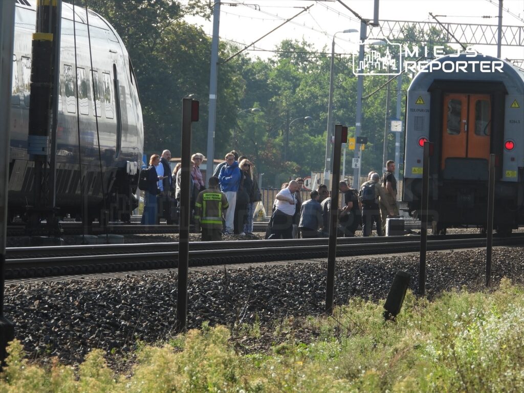 Grupa ludzi czeka obok torów kolejowych, podczas gdy pracownik w kamizelce informacyjnej rozmawia z pasażerami.