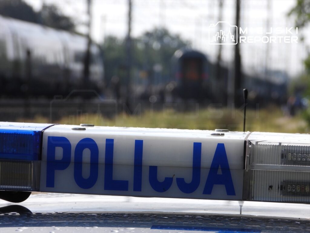 Policyjny radiowóz z niebieską sygnalizacją stoi na torach kolejowych, w tle widoczne pociągi.
