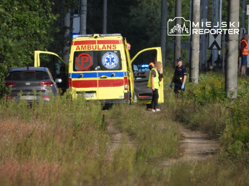 Ambulans z otwartymi drzwiami stoi obok drogi, a ratownicy i policjant rozmawiają w otoczeniu zieleni.