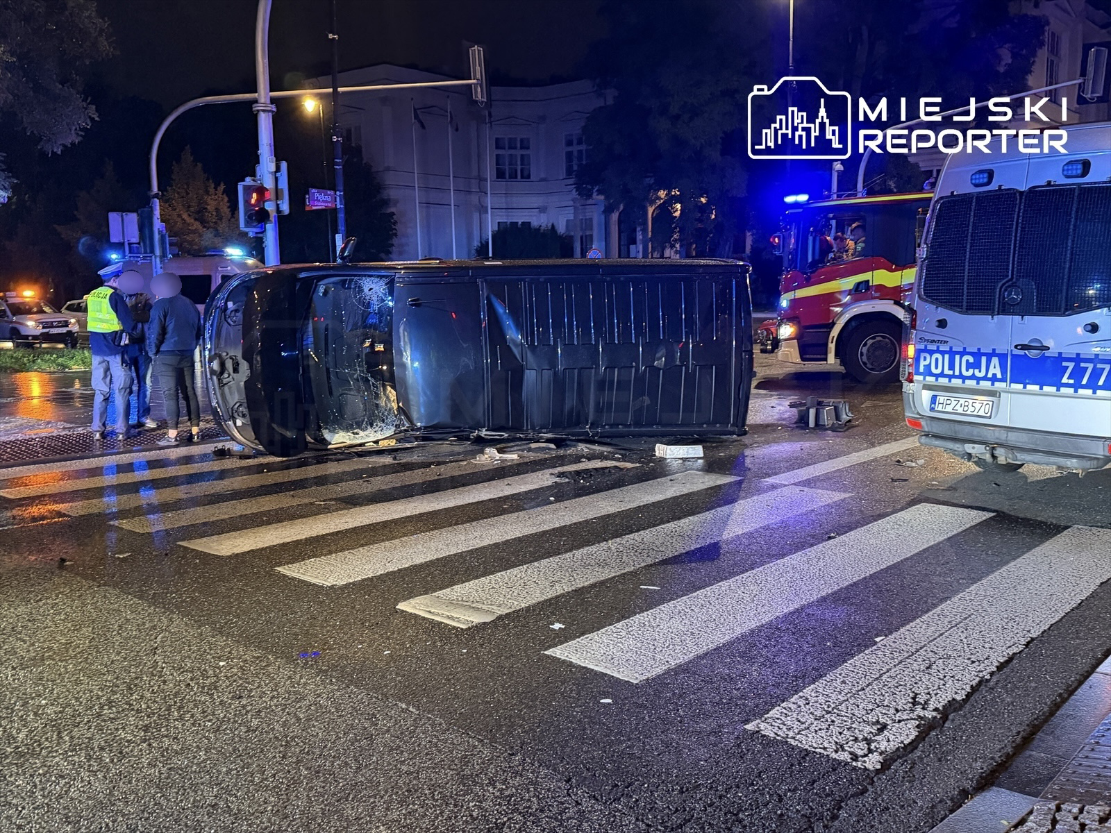Przewrócony samochód osobowy na pasach, obok policjanci rozmawiający z osobą, w tle woz strażacki.