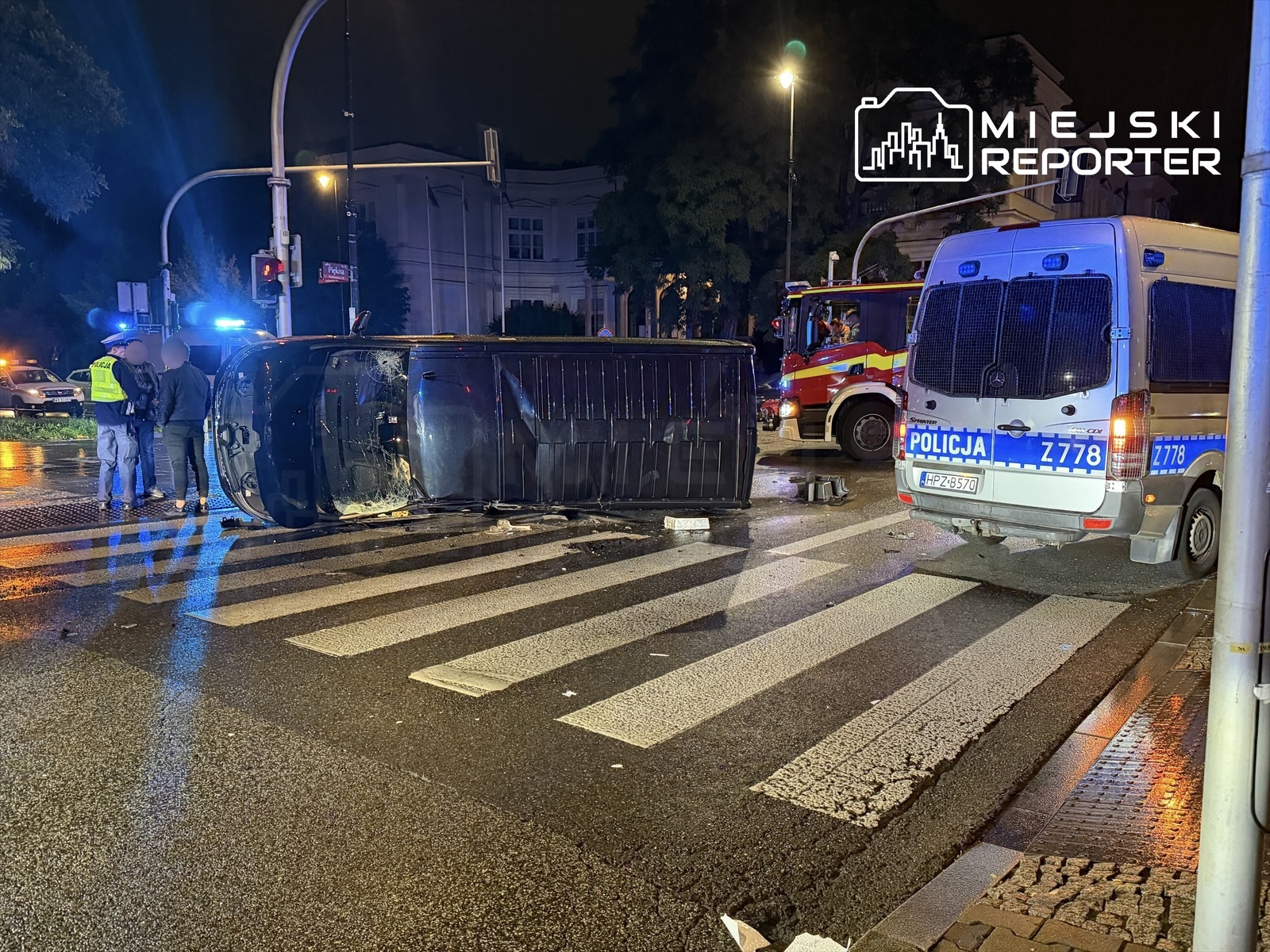Na pasach ruchu leży przewrócony samochód, obok stoi policjant rozmawiający z innymi osobami. W tle widać radiowóz i samochód strażacki.