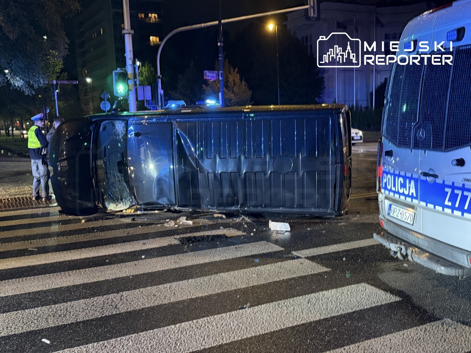 Nieoznakowany pojazd leży na boku na pasach, obok policjant w kamizelce odblaskowej bada sytuację, w tle radiowóz.