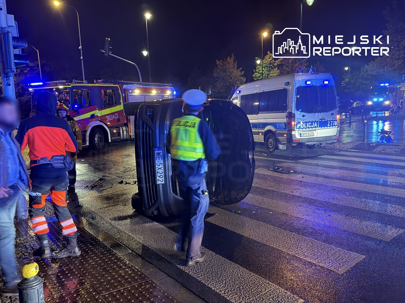 Na oświetlonej ulicy, funkcjonariusze policji oraz strażacy pracują przy przewróconym samochodzie osobowym, obok stoi wóz policyjny.
