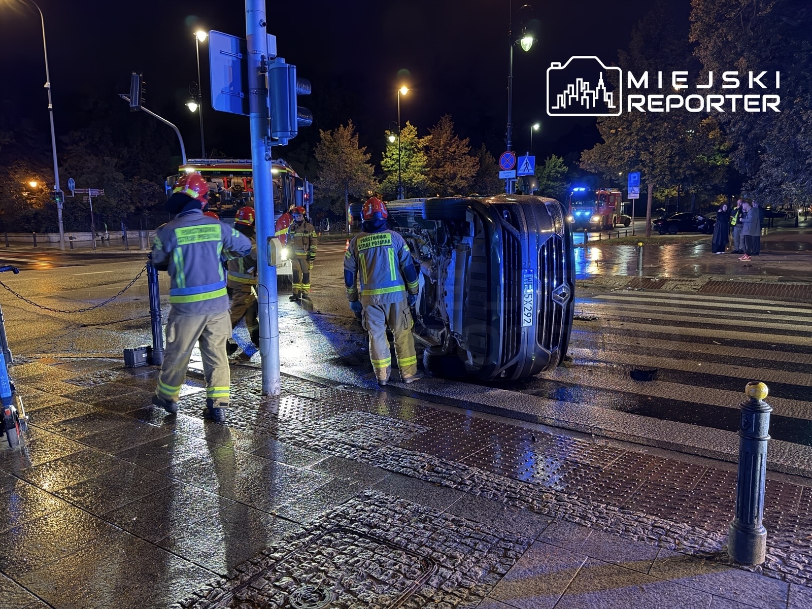 Grupa strażaków w mundurach i hełmach przemieszcza się przy przewróconym pojeździe na ulicy w nocy.