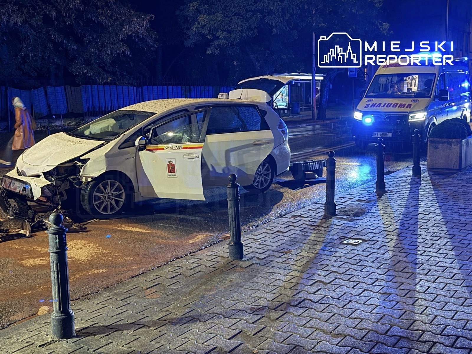 Rozbity taksówka z uszkodzoną przednią częścią stoi na jezdni, obok niej widać niebieskie światła karetki.