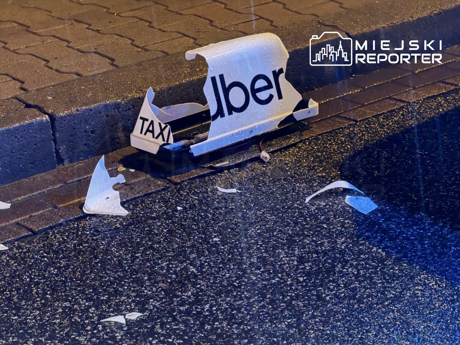Złamany znak taksówki z napisem "Uber" leży na mokrej nawierzchni obok krawężnika.
