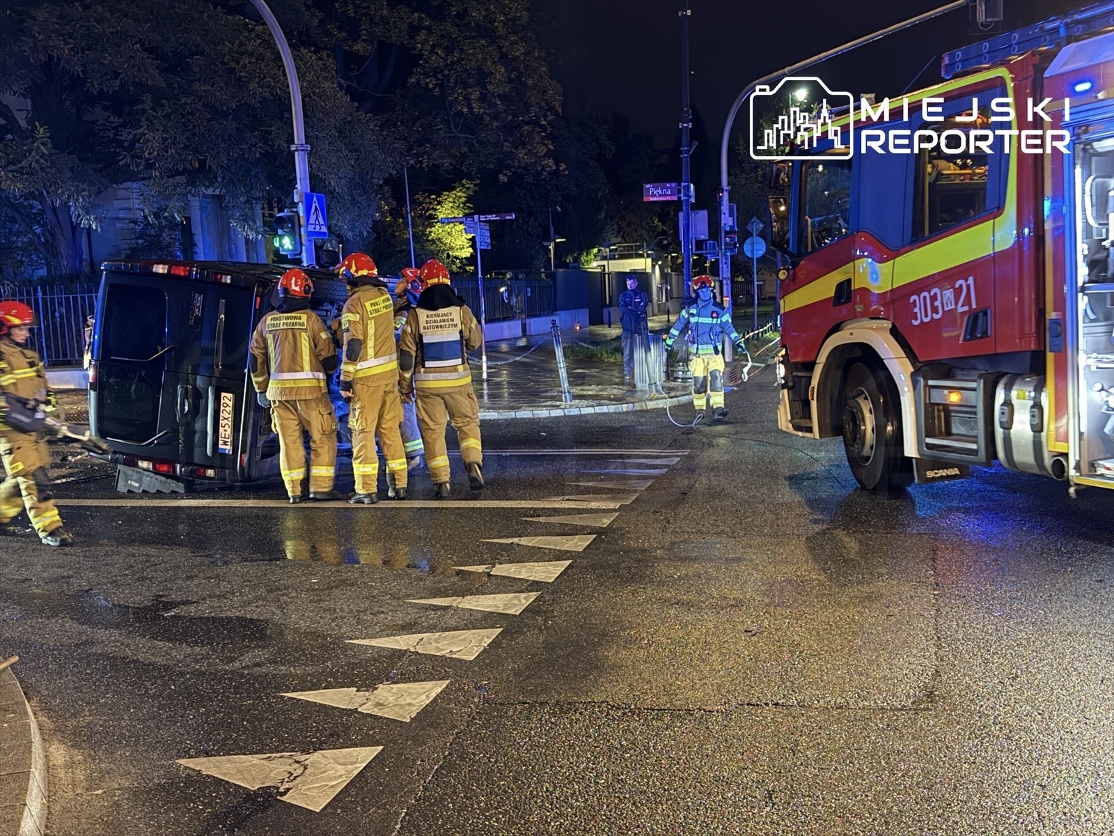 Grupa strażaków w hełmach bada przewrócony pojazd na mokrej ulicy w nocy, obok stoi wóz strażacki.
