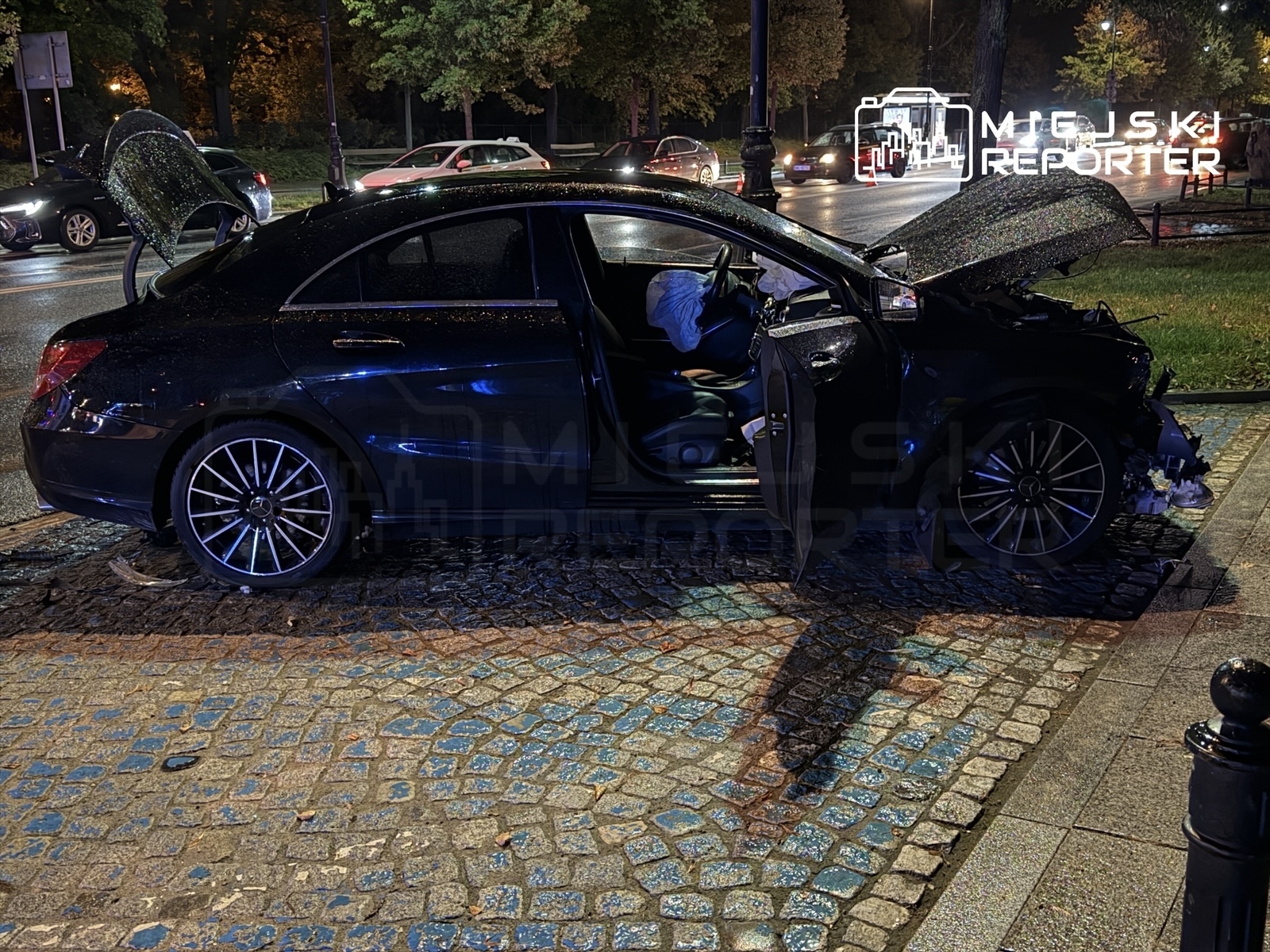 Czarny samochód osobowy z otwartą maską stoi na kostkowanej nawierzchni, obok widać inne pojazdy w tle.