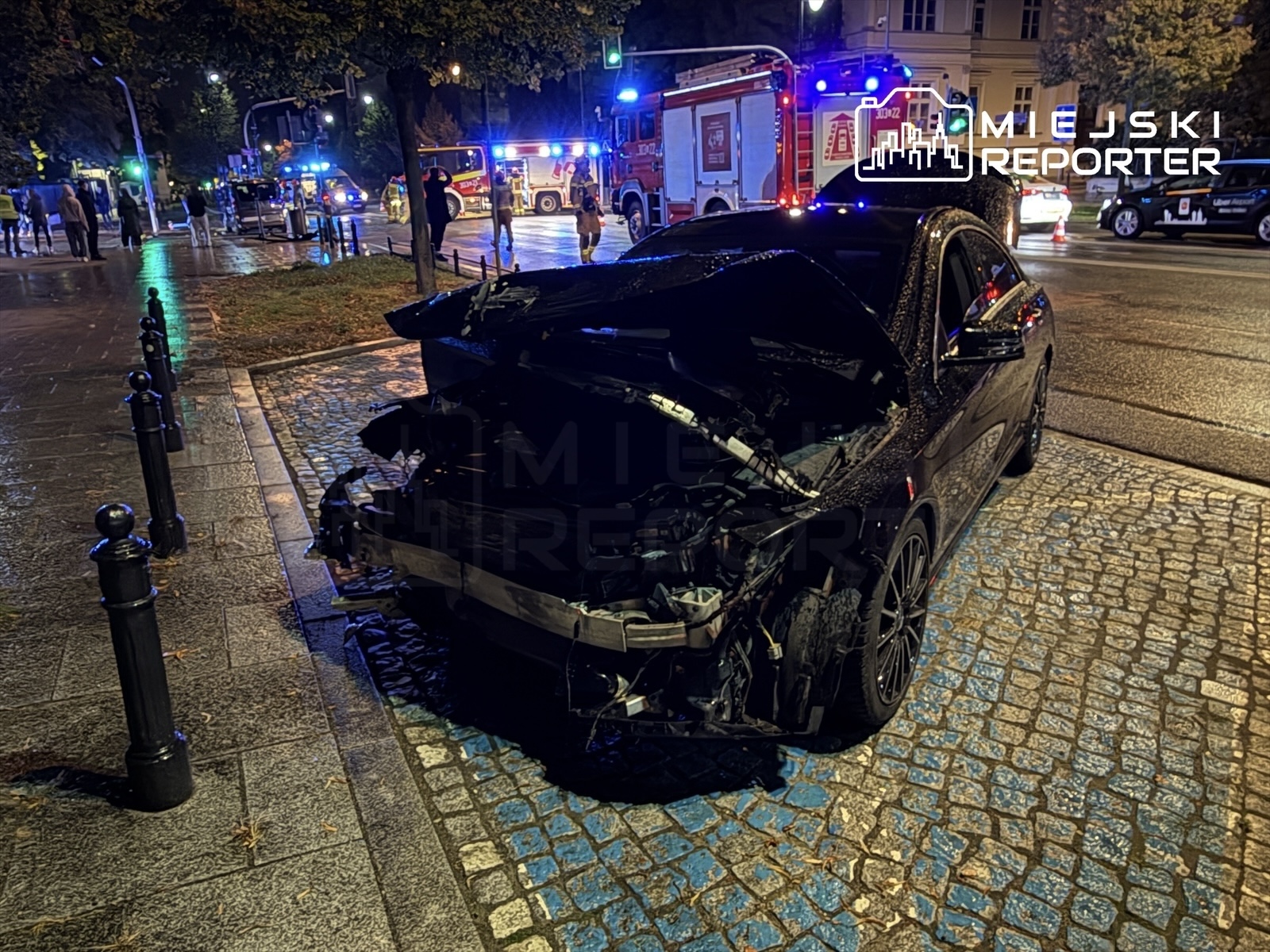 Uszkodzony czarny samochód osobowy stoi na chodniku, obok widać pojazdy służb ratunkowych oraz zebranych ludzi.