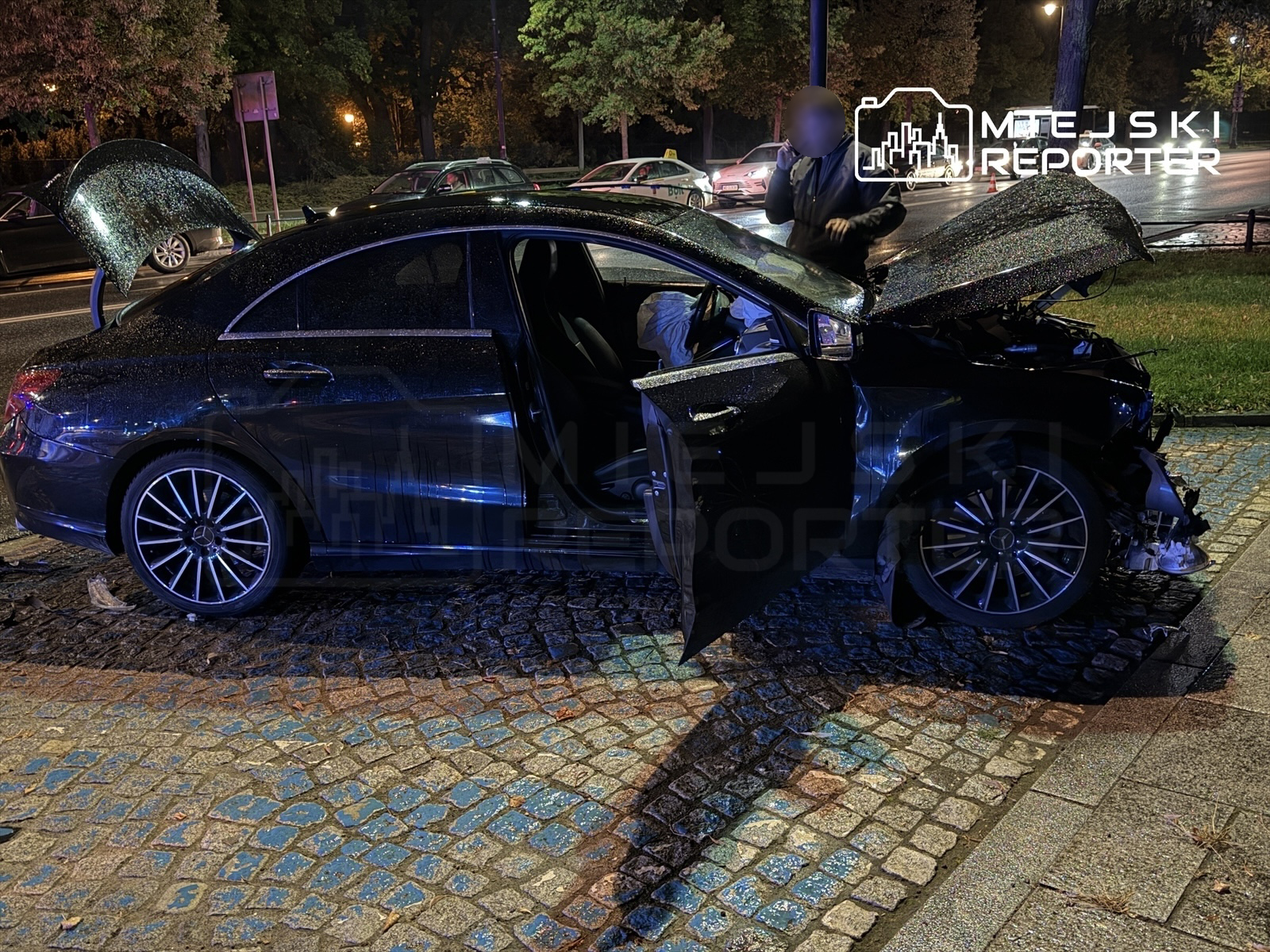 Czarny samochód osobowy z otwartą maską stoi na kostkowanej nawierzchni, obok mężczyzna rozmawia przez telefon.