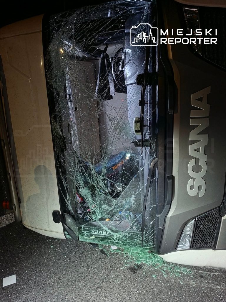Przewrócona cysterna marki Scania z rozbitym przednim szkłem leży na boku na drodze, wokół rozbite szkło.
